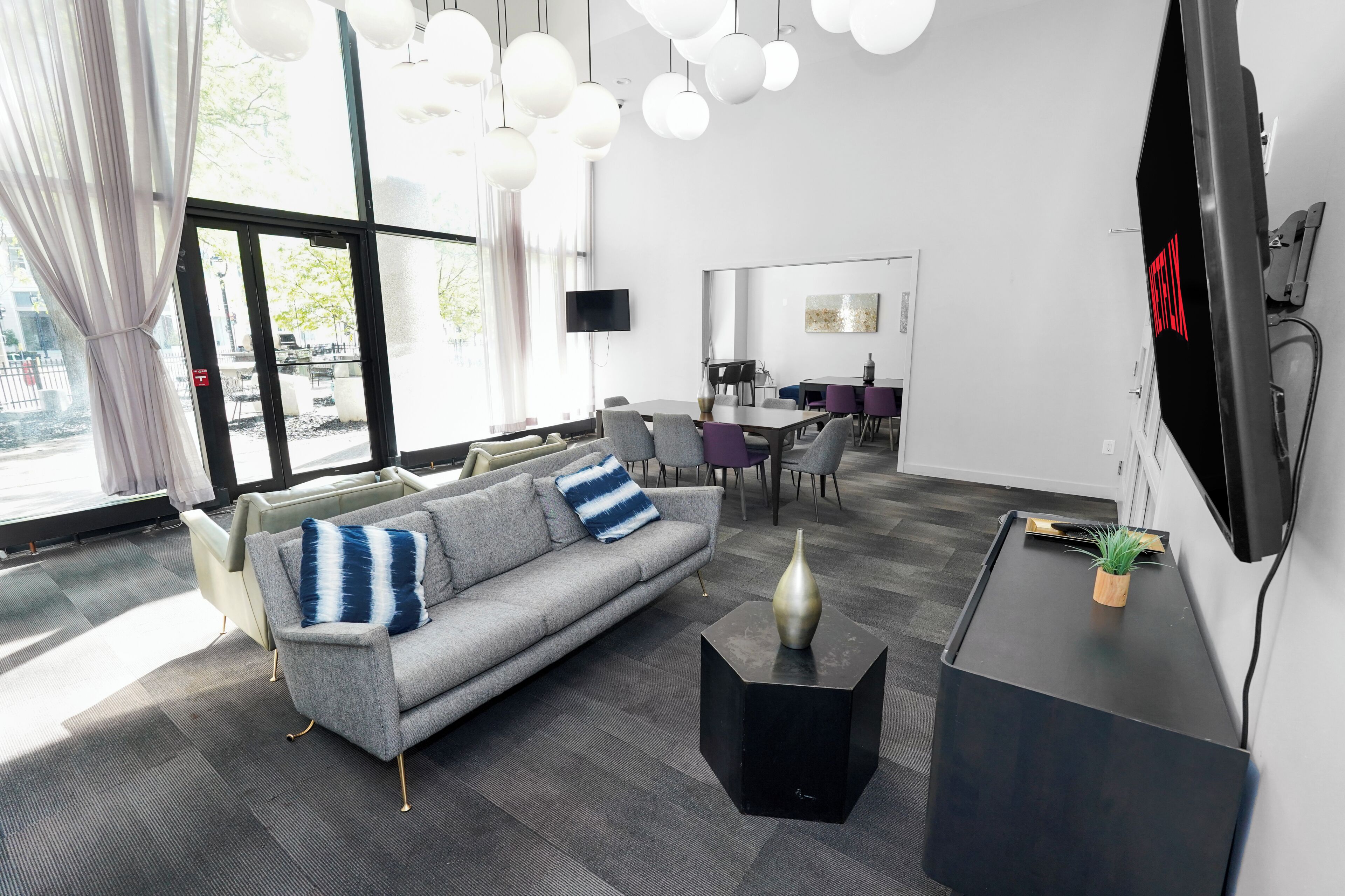 Habitació d'hotel moderna amb un sofà gris, una taula de cafè negra, una televisió i una zona de menjador al fons.