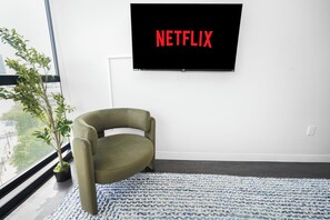 Et moderne værelse med et stort vindue, en potteplante, en sofa og et vægmonteret TV, der viser 'NETFLIX'.