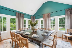 Uma sala de jantar com uma mesa grande posta para seis pessoas, rodeada por cadeiras de madeira, janelas amplas e vista para o exterior com vegetação.