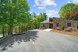 A modern stone house with a driveway, surrounded by trees and a clear sky.