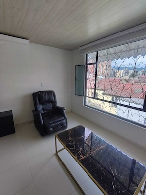 A room with a black leather chair, a glass-top coffee table, and a large window with metal bars.