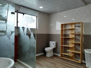 A bathroom with a glass shower enclosure, a toilet, a wooden shelf unit, and a white laundry basket.