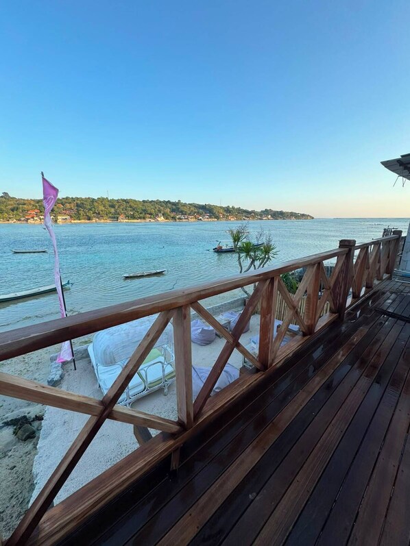 Una terraza de madera con vistas a una superficie acuática tranquila, con barcos, una orilla lejana y una bandera.