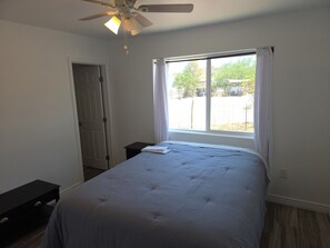 A bedroom with a bed, a ceiling fan, a window with curtains, a door, and a small table.