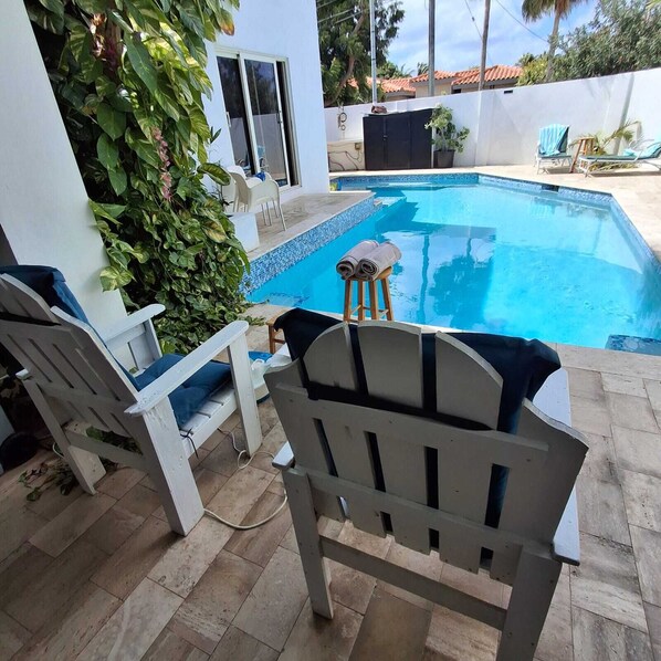 A swimming pool with lounge chairs and a small table, surrounded by a white wall and greenery. - Casa Bella Villas & Spa (Noord)