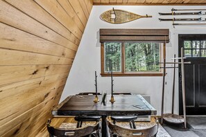 En inredning med träpanel, ett matbord, stolar, ett fönster med utsikt över träd och skidutrustning på väggen.