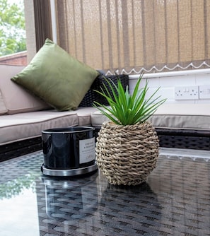 A wicker pot with a plant, a black scented candle, and a glass table in a room with a window and a sofa.