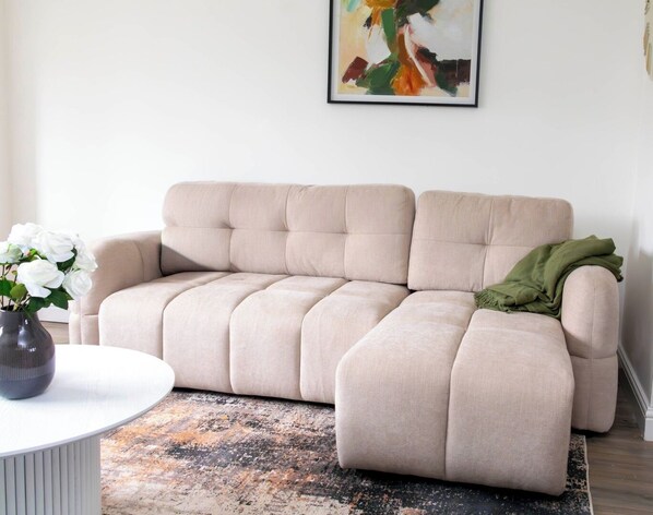 A beige sofa with a green throw blanket, a white vase with white flowers, and a painting on the wall.