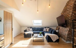 Ein Loft-Zimmer mit Schlafgalerie, einer Backsteinwand, einem an der Wand montierten Fernseher sowie einem kleinen Tisch mit Büchern und einer Pflanze.