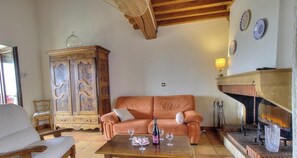 A living room with a sofa, a wooden cabinet, a fireplace, and a table with glasses and a bottle. - Le Camelia - CHENAS - Gîte (CHENAS)