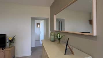 Un salon moderne comprenant une télévision, un meuble en bois, une commode beige, un grand miroir et un vase avec des fleurs.