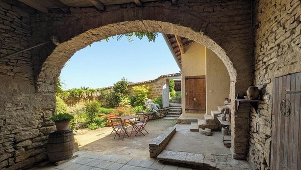 A stone archway leading to a patio with a table and chairs, surrounded by stone walls and a door. - Bird Paradise (POMMIERS)