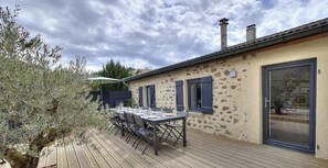 Eine Terrasse mit Esstisch, ein Steinhaus mit Fensterläden und eine Tür mit Glasfenster.