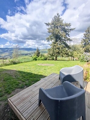 Eine Holzterrasse mit zwei Stühlen und einem Tisch, mit Blick auf eine Wiese mit Bäumen und einem fernen Gebirge.