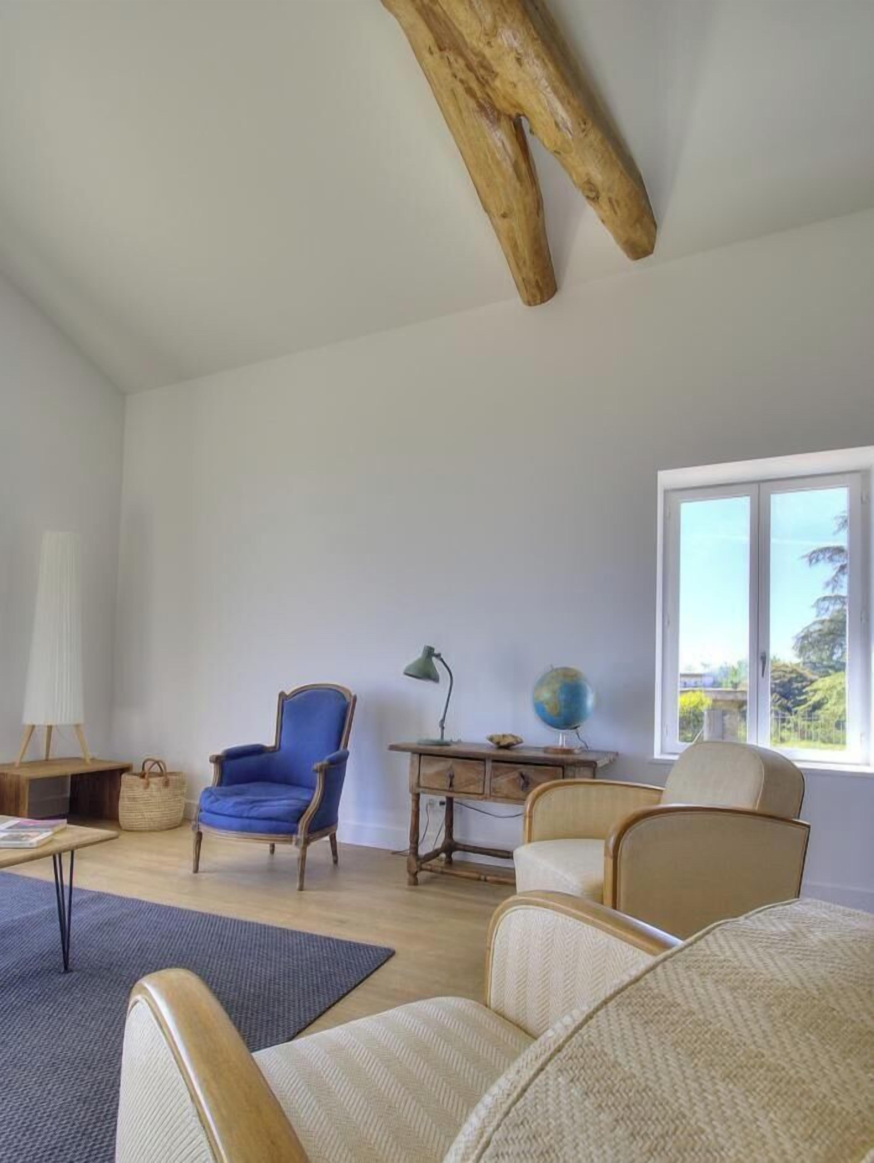 Un salón amplio con techo alto, un ventanal grande, un sillón azul, un sofá color beige y un escritorio de madera con un globo terráqueo.