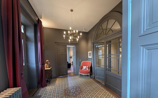 A hallway with patterned flooring, a chandelier, a red chair, and a door with glass panels.