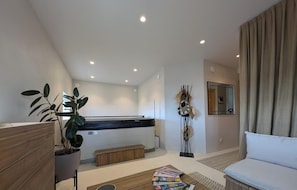 A modern living room with a sofa, a coffee table, a plant, a small bench, and a kitchen area with a white countertop and cabinets.