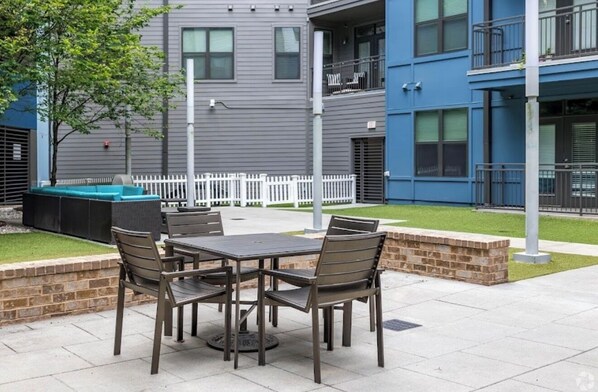 Une terrasse extérieure avec une table et des chaises, une clôture blanche et un bâtiment avec des balcons en arrière-plan.