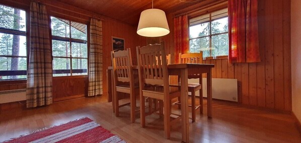 Ein Holzinterieur mit Esstisch, Stühlen, einer Hängelampe und großen Fenstern mit Vorhängen.