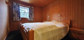 Ein Zimmer mit Holzwänden, einem Einzelbett, einem Nachttisch und einem fenster mit Vorhängen.