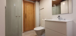 A bathroom with a glass shower enclosure, a wooden door, a white toilet, and a white sink with a mirror above it.
