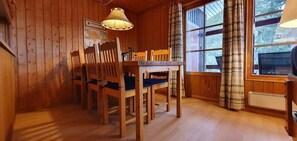 A wooden interior with a dining table and chairs, a hanging lamp, and a window with curtains.