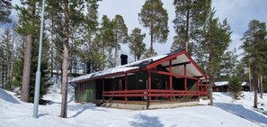 A cabin with a red exterior, surrounded by snow-covered ground and tall pine trees.