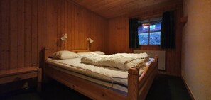 A wooden cabin room with a single bed, a window with curtains, and a wall-mounted lamp.