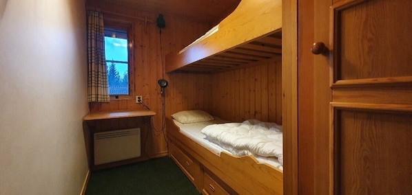 Ein kleines Zimmer mit Holzwänden, ausgestattet mit einem Etagenbett, einem Schreibtisch mit Lampe und einem Vorhangfenster.