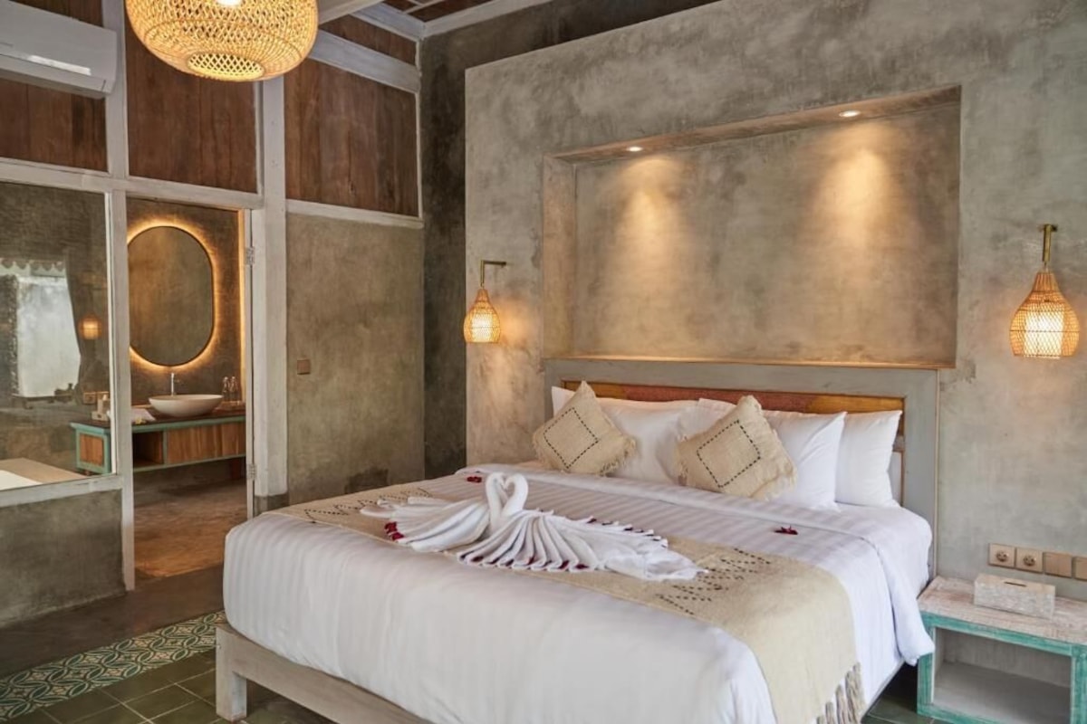 Ein Schlafzimmer mit einem Bett, Nachttischen, einer Wandleuchte und einem Badezimmer mit Waschbecken und Spiegel.