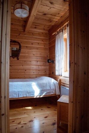 Ein Zimmer in einer Holzhütte mit einem Bett, einem kleinen Tisch, einem Fenster mit Vorhängen und einer Pendelleuchte.
