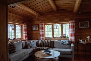 A wooden cabin interior with a sofa, a round table, and windows with curtains.