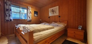 Ein Zimmer aus Holz mit einem Bett, einem Nachttisch, einem Fenster mit Vorhängen und einem gerahmten Bild an der Wand.