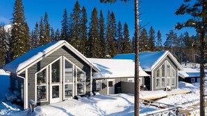 En tvåvåningsstuga med stora fönster och en inglasad veranda, omgiven av snö och tallar.