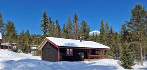 Une cabane avec un toit rouge et une véranda, entourée de neige et d’arbres.