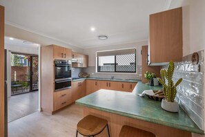 A modern kitchen with wooden cabinets, a built-in oven, and a large window. - Coastal family home - 4BR, walk to beach (Warana)
