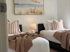Ein Hotelzimmer mit zwei Betten, gestreiften Kissen, einem Meeresmotiv-Bild, einer Lampe und einer Decke.