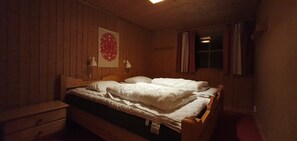 Ein Zimmer mit Holzvertäfelung, einem Einzelbett, einem Nachttisch, einer Wandlampe und einem mit Vorhängen drapierten Fenster.