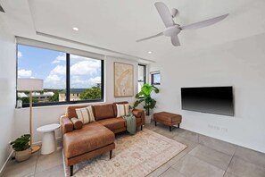 Ein modernes Wohnzimmer mit einem Ecksofa, einem Deckenventilator, einem großen Fenster mit Blick auf die Stadt, einem Flachbildfernseher und einer Topfpflanze.