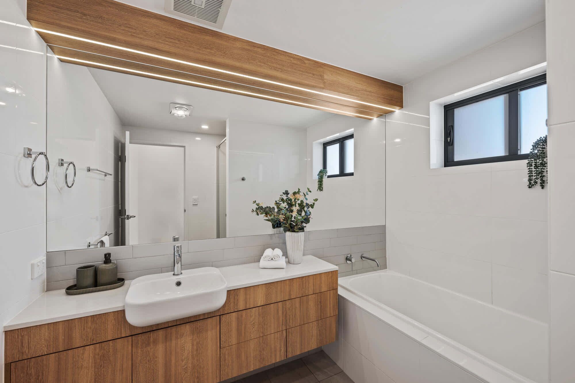 Ein modernes Badezimmer mit einem Holzkosmetikschrank, einem großen Spiegel, einer Badewanne und einem kleinen Fenster.