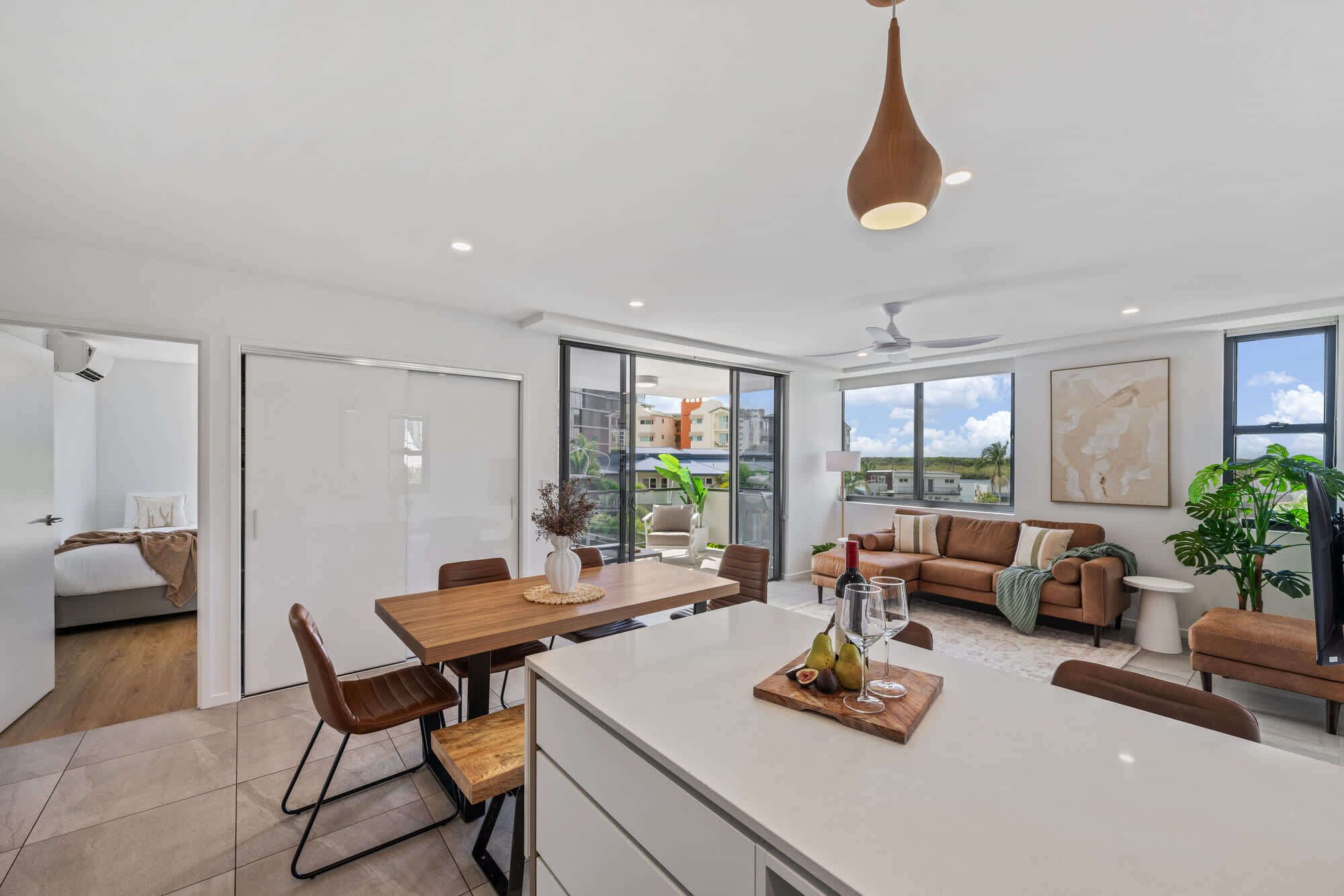 A modern kitchen with a central island, dining area, and a living space with a sofa and a large window.