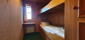 Ein kleiner, holzgetäfelter Raum im Blockhausstil mit einem Stockbett, einem kleinen Schreibtisch mit Stuhl und einem Fenster mit Vorhängen.