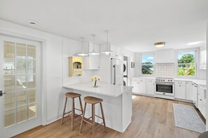 A modern kitchen with white cabinetry, a central island with stools, a built-in oven, and a large window offering a view of greenery.