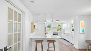 A modern kitchen with white cabinetry, a central island with stools, pendant lighting, and a view of greenery outside. - Newly Renovated Clinton Beach House (Clinton)