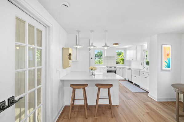 Une cuisine moderne avec des meubles blancs, un îlot central doté de tabourets, un éclairage suspendu et une vue sur la verdure à l’extérieur.