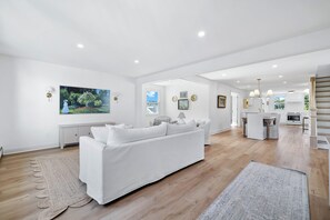 A spacious living area with a white sofa, a kitchen in the background, and a view of the outdoors.