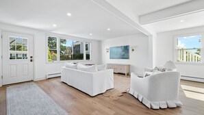 A spacious living room with a white sofa, a white armchair, a television, and large windows. - Newly Renovated Clinton Beach House (Clinton)