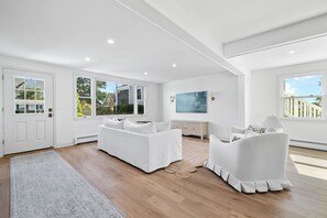A spacious living room with a white sofa, a white armchair, a wooden floor, and large windows.