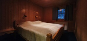 Ein Zimmer mit Holzwänden, einem Einzelbett, einem Nachttisch, einer Lampe und einem Fenster mit Vorhängen.