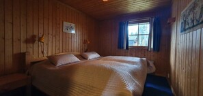 Ein Zimmer in einer Holzhütte mit einem Bett, einem Fenster mit Vorhängen, einer Wandlampe und einem gerahmten Bild.